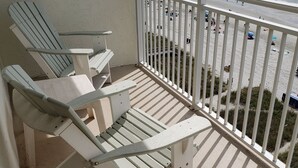 Ein Balkon mit weißen Stühlen und einem kleinen Tisch mit Blick auf einen Strand, auf dem sich Menschen und Sonnenschirme befinden.
