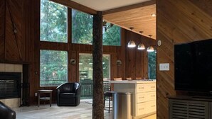 A spacious living room with a fireplace, a large window, a kitchen area, and a television. - Stellar Blue Chalet - Sleeps 6+. Pets on approval. Near Alyeska Ski Resort (Girdwood)