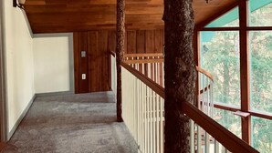 A wooden interior hallway with a carpeted floor, a wooden railing, and a view of trees through large windows. - Stellar Blue Chalet - Sleeps 6+. Pets on approval. Near Alyeska Ski Resort (Girdwood)