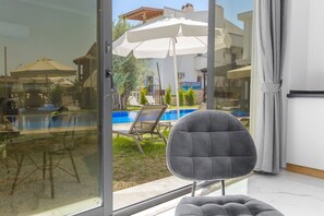 A modern room with a glass door leading to a pool area, a grey cushioned chair, and a white umbrella. - Nur Suites & Hotels (Kas)