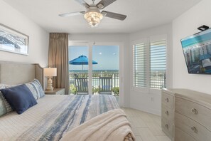 A bedroom with a bed, a ceiling fan, a window with blinds, a balcony with a blue umbrella, and a TV on the wall. - Blu 107 ~ Oceanfront 4BR/3BA ~ Sleeps 10 ~ Ground Floor End Unit! (Fort Walton Beach)