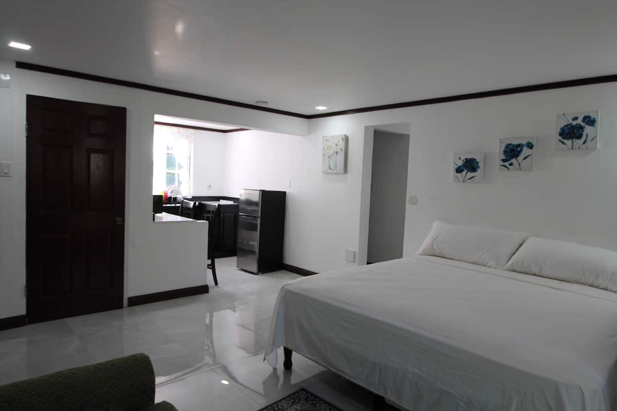 A bedroom with a large bed, a desk, and a kitchenette in the background.