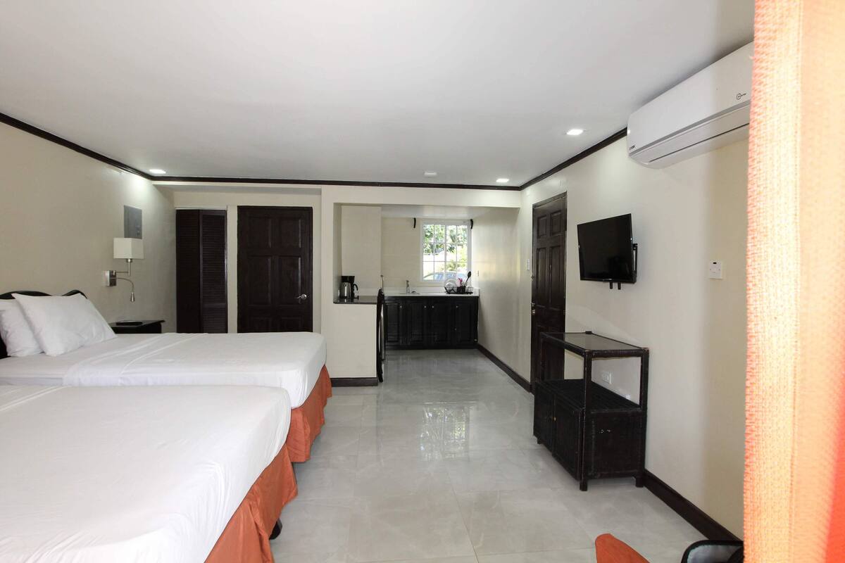A hotel room with two beds, a kitchenette, a TV, and an air conditioning unit.