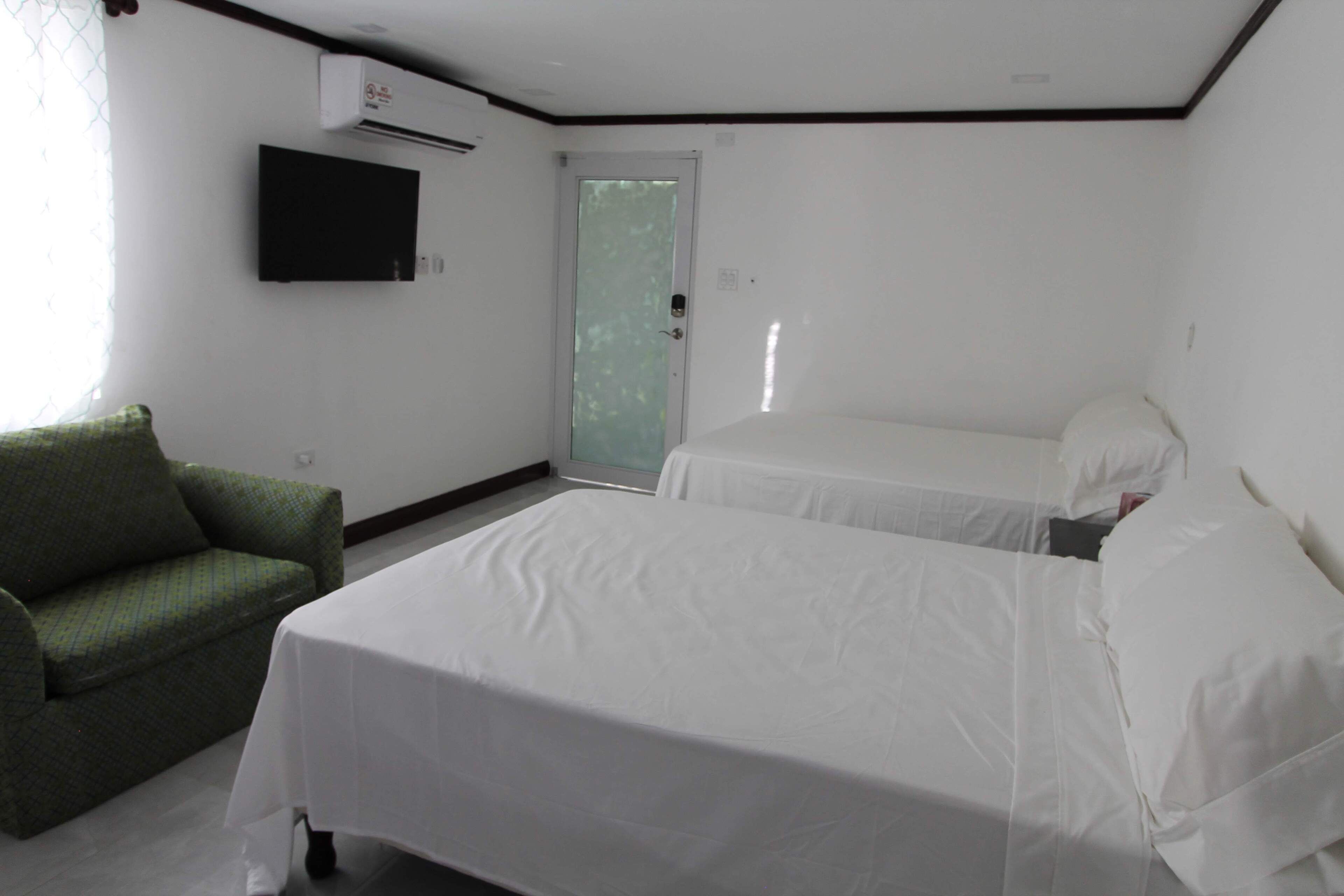 A hotel room with two beds, a green armchair, a flat-screen TV, and an air conditioning unit.
