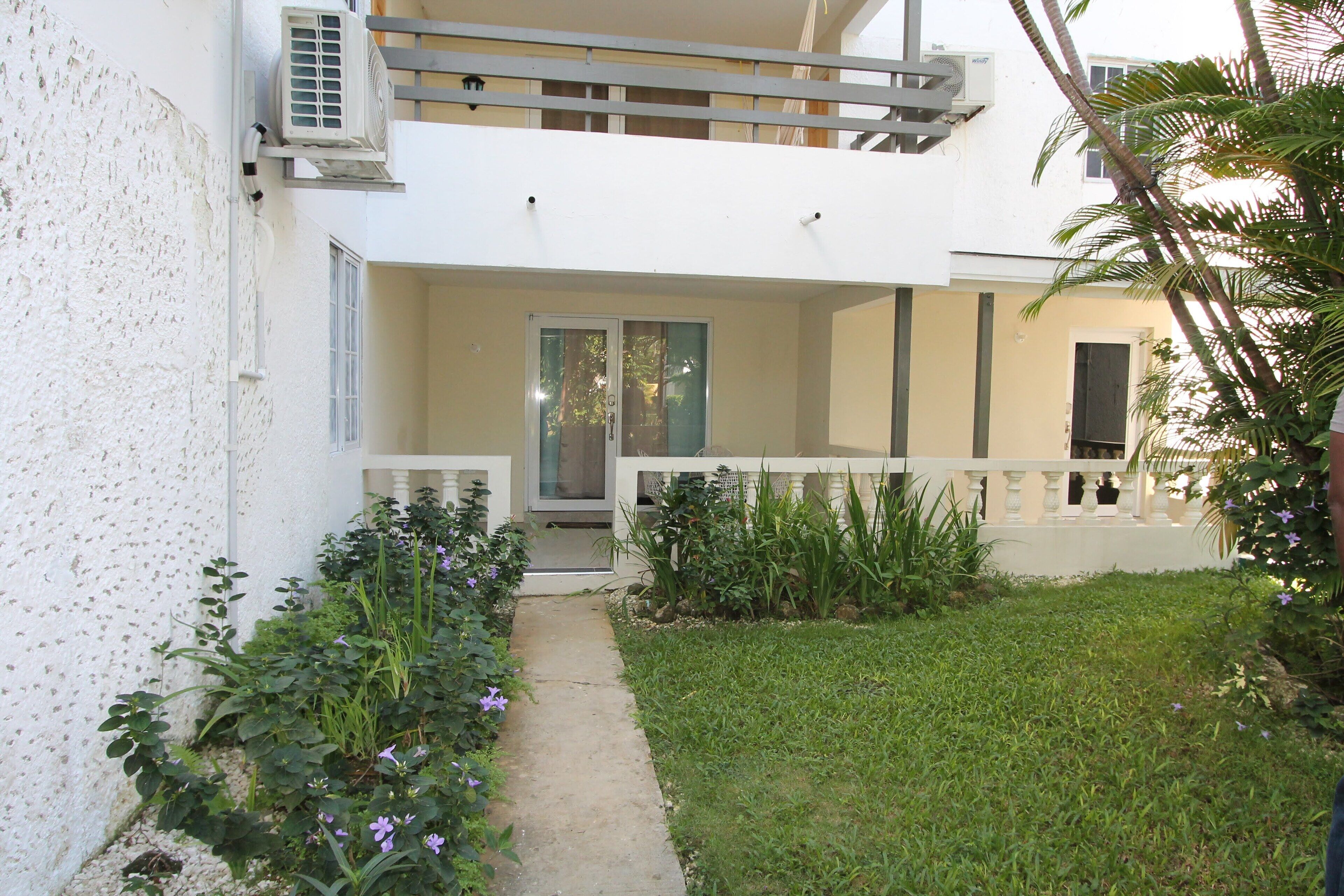 A two-story building with a balcony, a door with glass panels, and a pathway leading to the entrance.