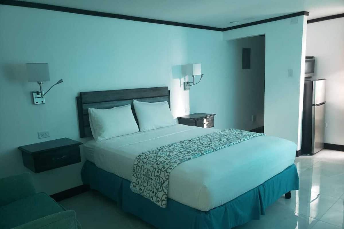 A hotel room with a bed, bedside tables, a nightstand, a chair, and a refrigerator.