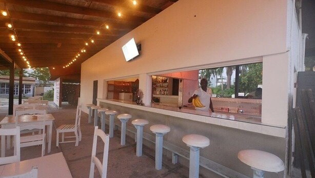 An outdoor seating area with white tables and stools, a bar counter, and a television mounted on the wall. - Sand and Blue Seas (Negril)