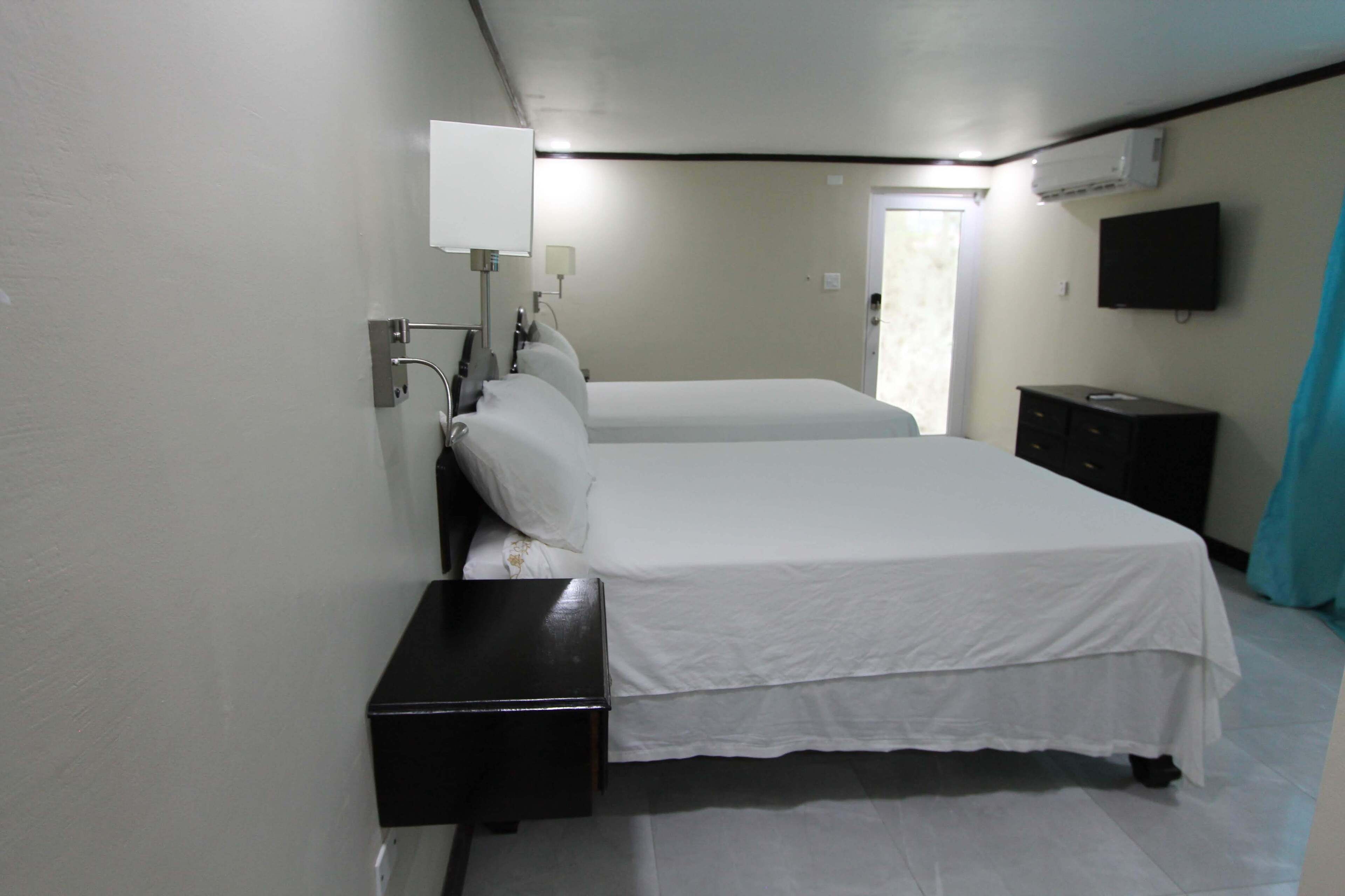 A hotel room with two beds, a bedside table, a TV mounted on the wall, and a window with curtains.