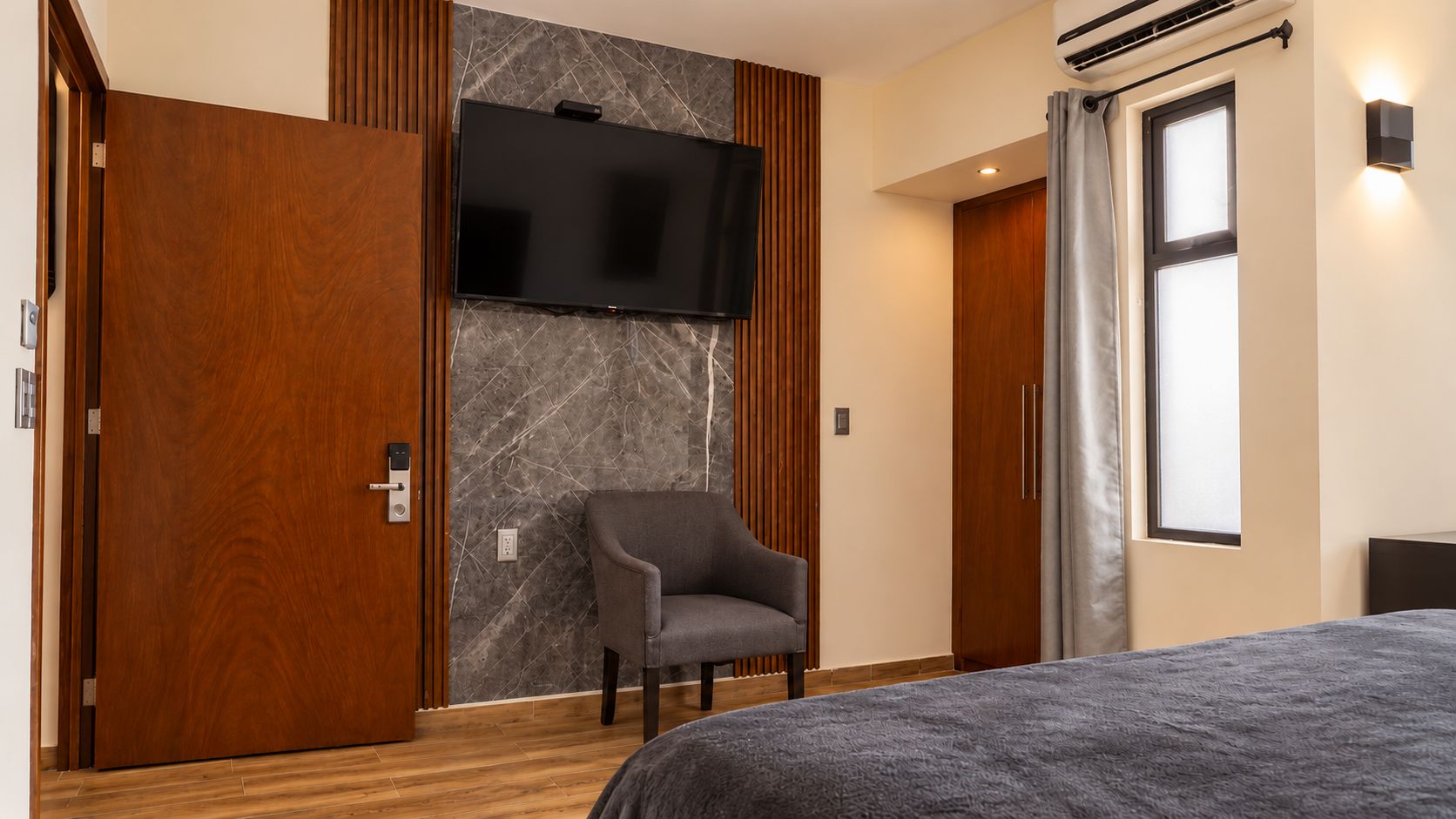 Una habitación de hotel moderna con una cama grande, un televisor de pantalla plana montado en una pared de mármol, una puerta de madera y una ventana con cortinas.