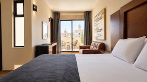 Een hotelkamer met een groot bed, een bureau, een stoel en uitzicht op een stadsgezicht door een raam.