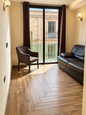 Une pièce avec un plancher en bois, un canapé en cuir marron, un fauteuil marron et une porte-fenêtre coulissante donnant sur un balcon.