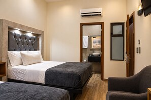 Une chambre d’hôtel comprenant un lit, deux tables de chevet, une chaise, une salle de bain avec un lavabo et un miroir, et un appareil de climatisation.
