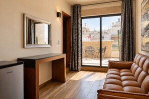 Ein möbliertes Zimmer mit Schreibtisch, einer Couch, einem großen Fenster mit Vorhängen und Blick auf Gebäude.