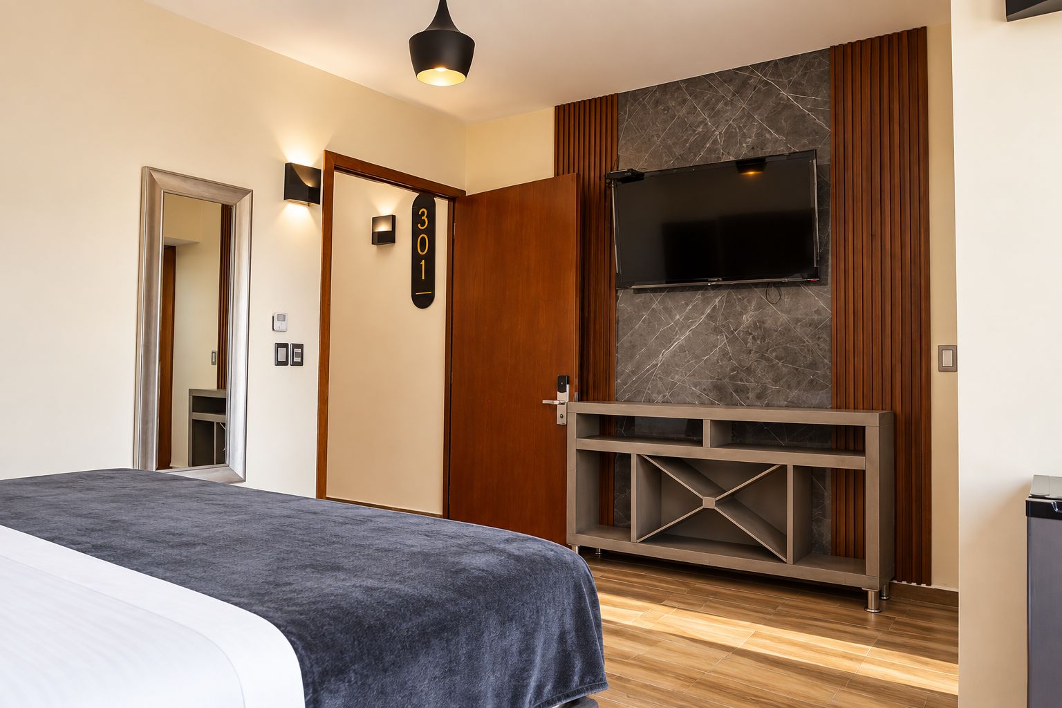 Ett hotellrum med en säng, en tv som är monterad på väggen, en trädörr och ett litet skåp.