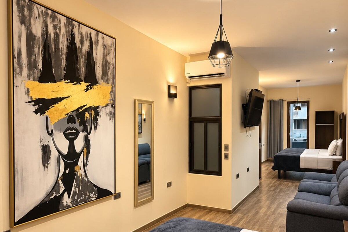 Une chambre d’hôtel moderne avec un grand tableau abstrait, un lit recouvert d’un plaid bleu foncé, un canapé, une télévision et un couloir menant à un balcon.