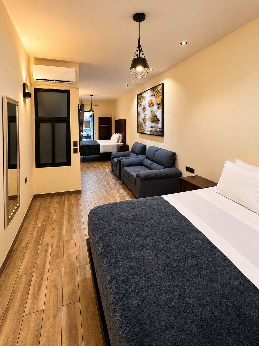 A modern hotel room with a large bed, a sofa, a desk, and a view into the dining area.