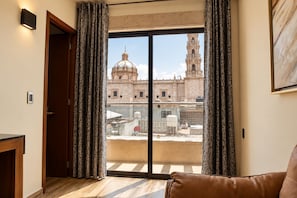 Ein Zimmer mit einer großen Schiebeglasdoor, die auf einen Balkon führt, einem braunen Ledersofa und Blick auf ein historisches Gebäude mit Kuppel und Glockenturm.