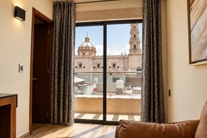 Una habitación con un gran ventanal corredero que da a un balcón, un sofá de cuero marrón y vistas a un edificio histórico con cúpula y campanario.