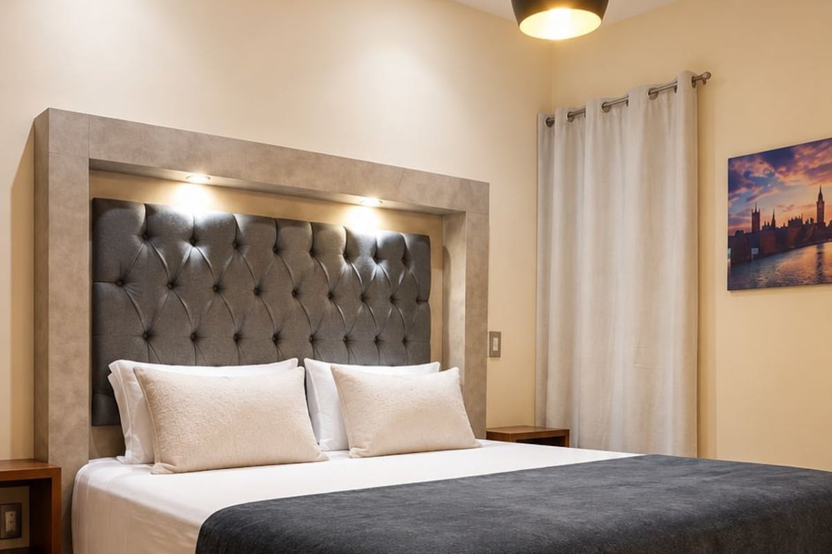 Une chambre d’hôtel moderne avec un grand lit, une tête de lit capitonnée, une couvre-lit de couleur sombre, un plancher en bois, une table de chevet et une lampe suspendue.