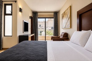 Ein Hotelzimmer mit Bett, Fernseher, Schreibtisch, Stuhl und einem Balkon mit Blick auf Gebäude.
