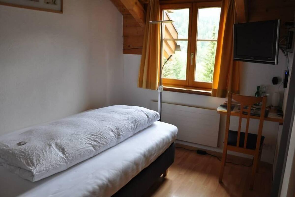 A small, single-bed room with a wooden desk and chair, a window with curtains, and a television mounted on the wall.