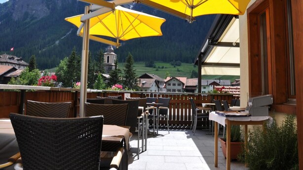An outdoor patio with yellow umbrellas, wicker chairs, and a view of a mountain village. - Hotel Ladina (Berguen)