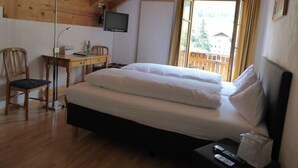 A bedroom with a wooden ceiling, a bed with white bedding, a desk with a chair, a TV mounted on the wall, and a window with curtains. - Hotel Ladina (Berguen)