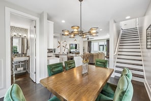 A modern kitchen with white cabinets and a central island, a dining area with a wooden table and green chairs, and a staircase with a railing.