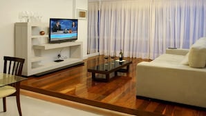 A modern living room with a wooden floor, a glass coffee table, a white sofa, and a television on a white stand. - Ipanema (Rio de Janeiro)