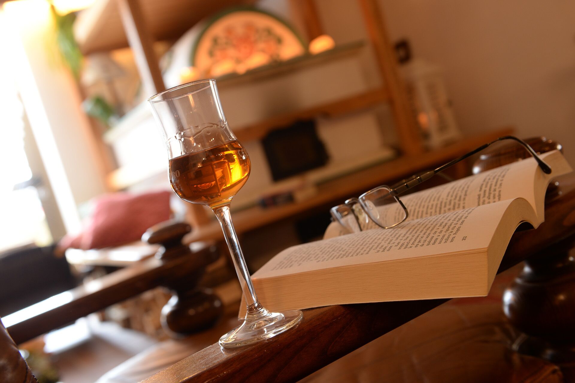 A glass of amber liquid on a wooden surface with a book and glasses beside it.