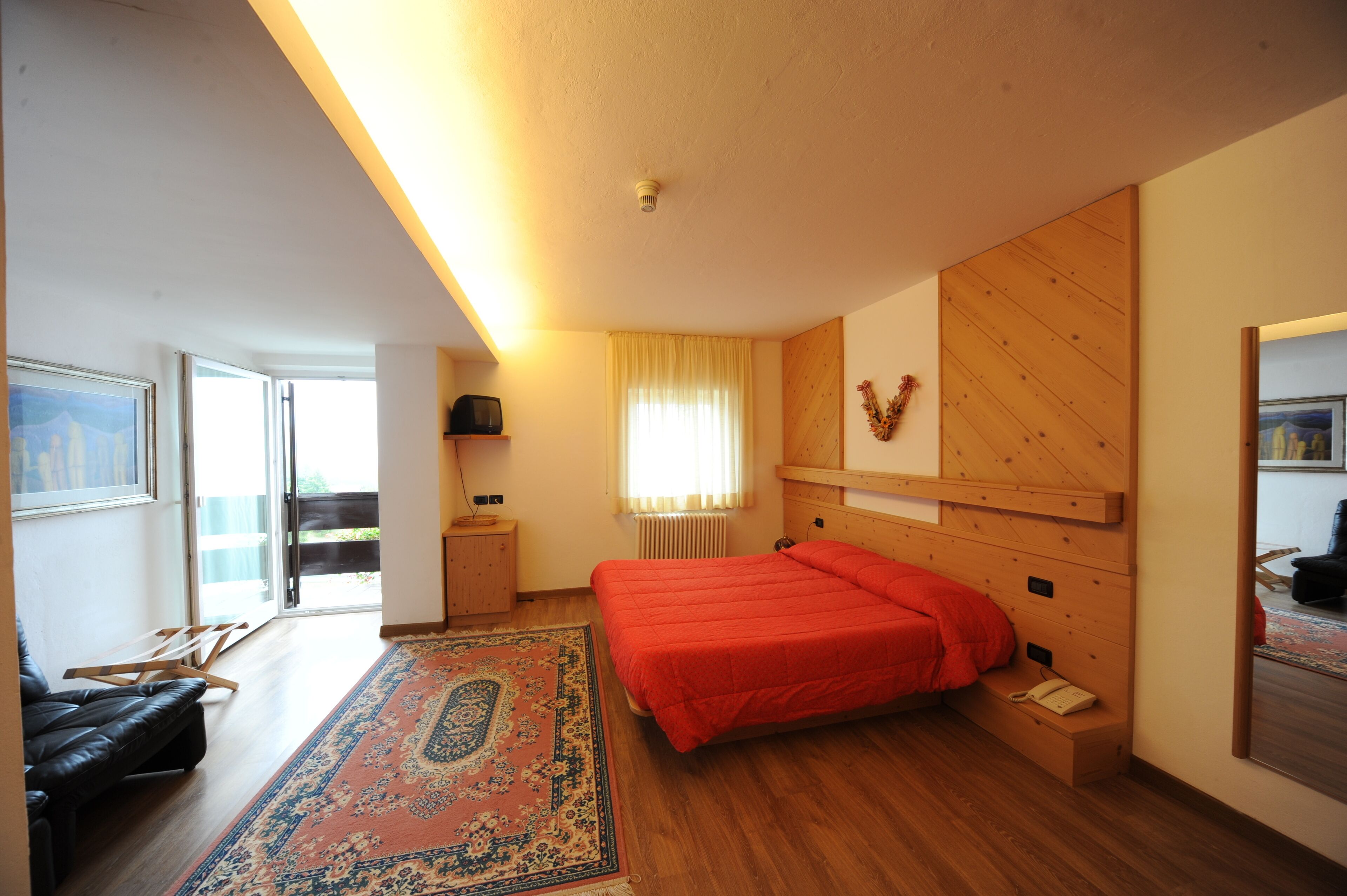 Un dormitorio con cama, cabecera de madera, mesita de noche, una silla, ventana con cortinas y un radiador.