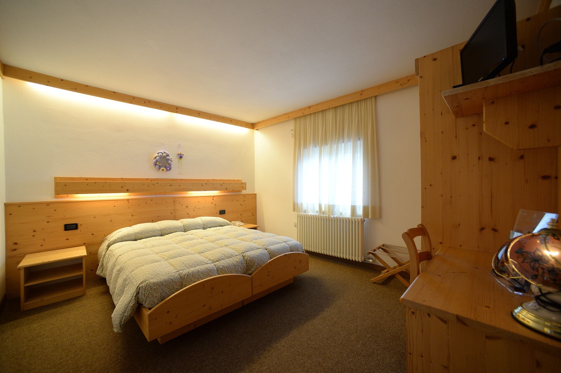 A wooden bedroom with a bed, a bedside table, a chair, a radiator, and a television on a wooden stand.