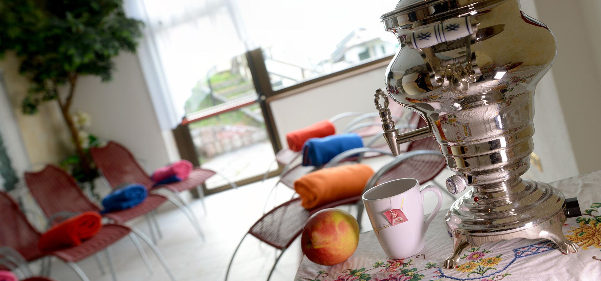 Een tafel met een zilveren theepot, een roze mok en een perzik. Op de achtergrond staan stoelen met kleurrijke kussens.