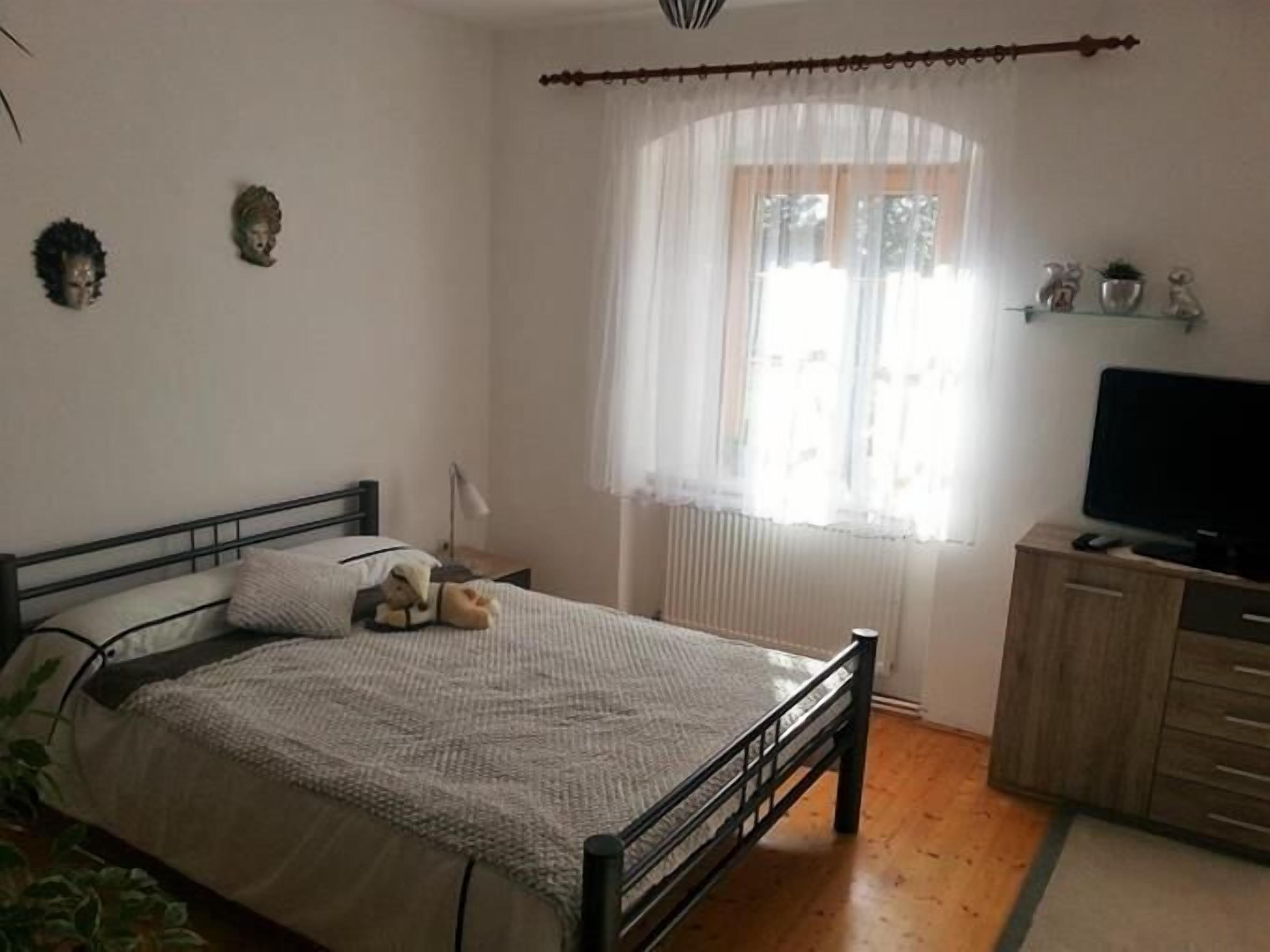 A bedroom with a bed, a bedside table, a lamp, a TV, and a window with curtains.