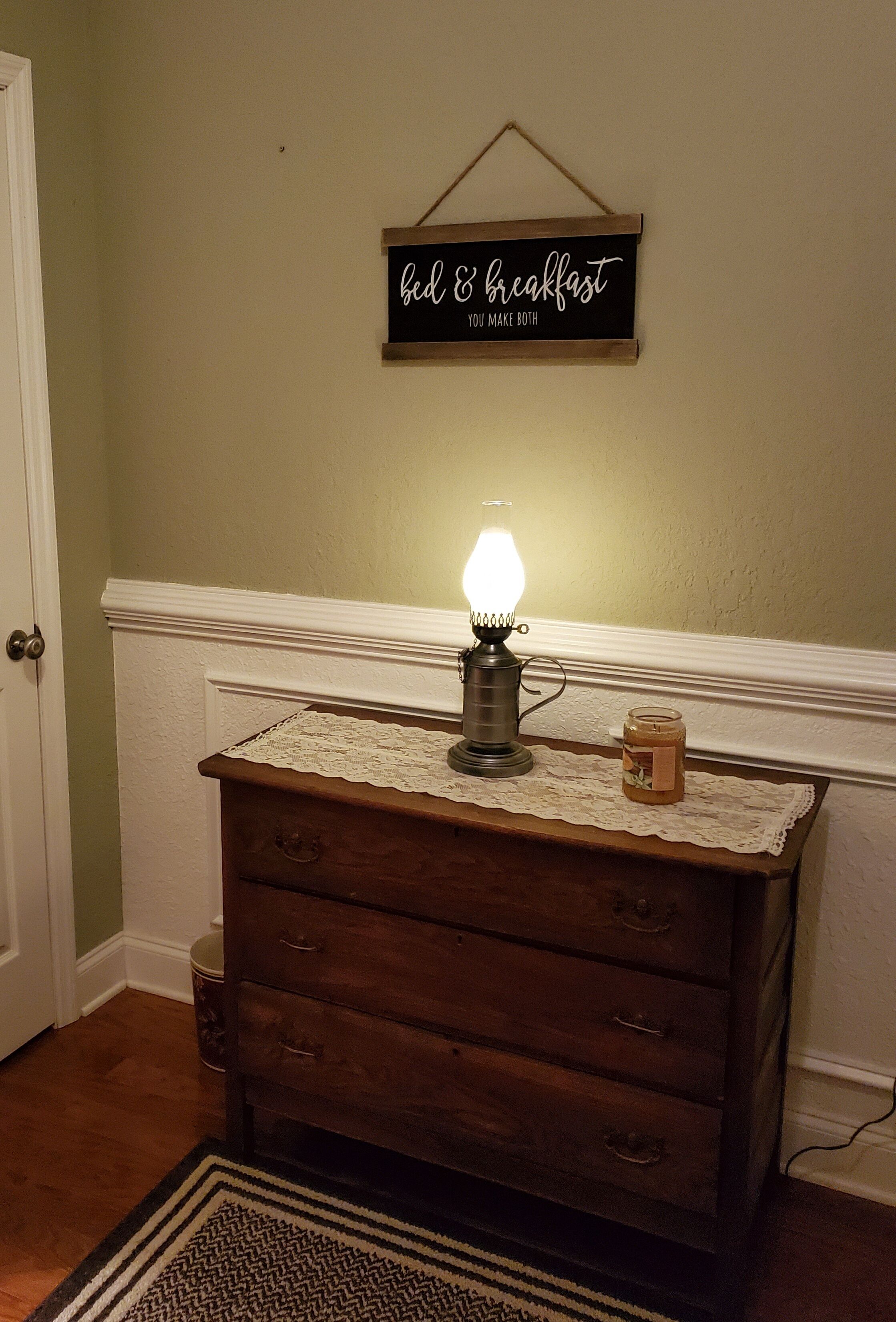 A wooden dresser with a lit lamp, a jar of honey, and a sign on the wall that reads "bed & breakfast you make both."