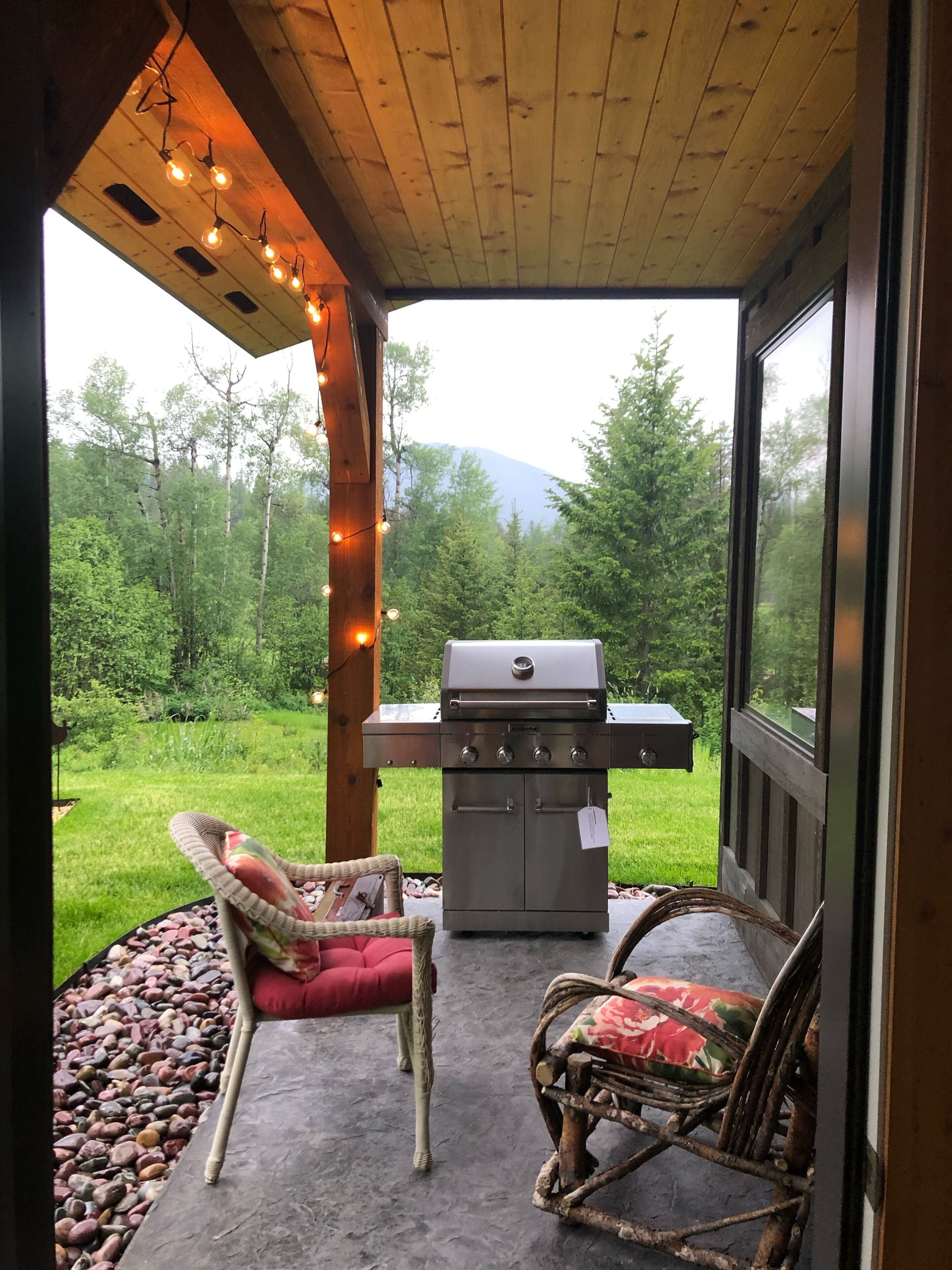Une terrasse extérieure avec un barbecue, deux fauteuils en osier avec des coussins et une guirlande lumineuse.