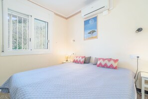 Un dormitorio con cama, mesita de noche, unidad de aire acondicionado, ventana y un cuadro colgado en la pared.