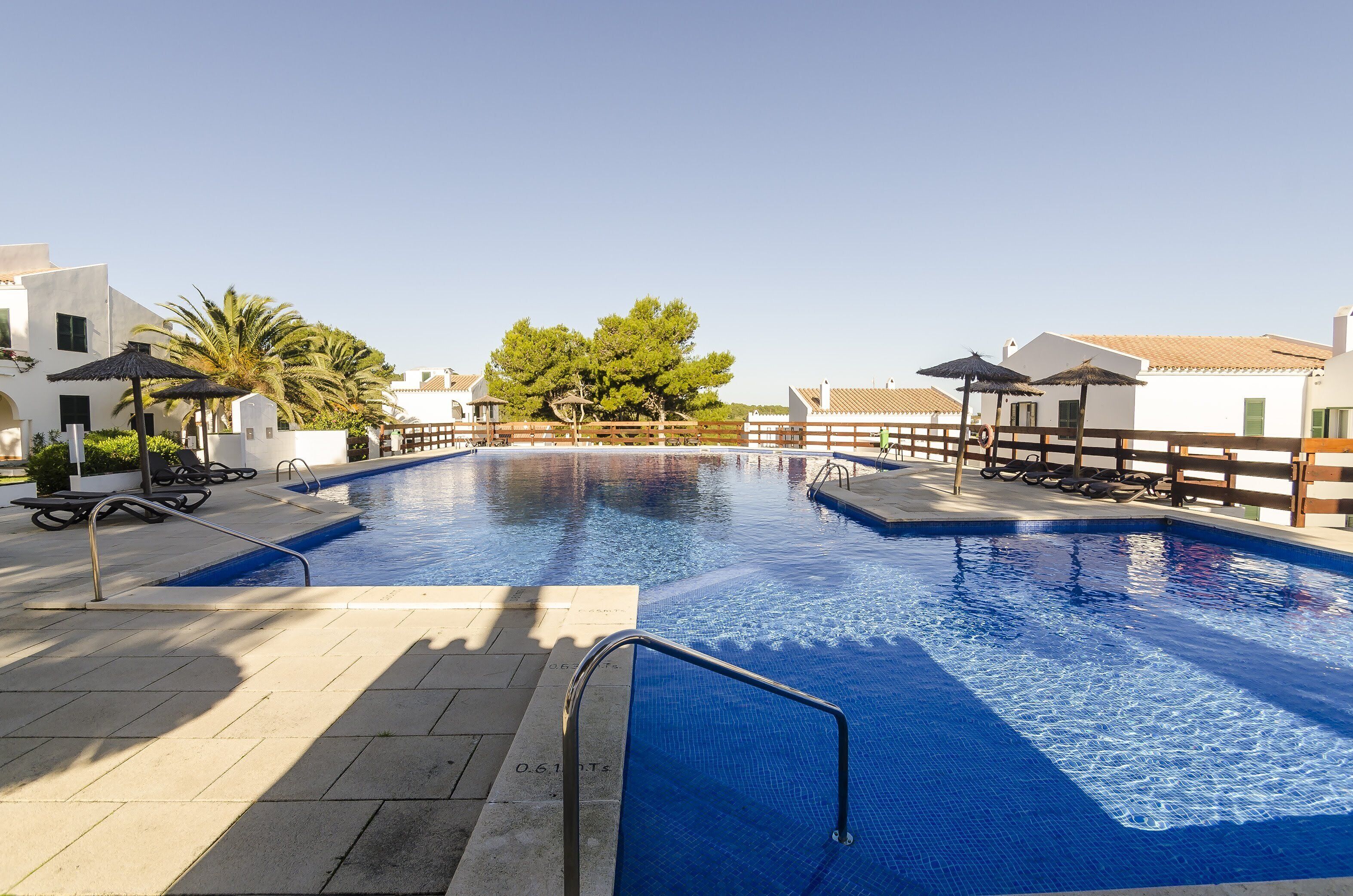 Una piscina con agua azul cristalina, rodeada por una terraza de madera, palmeras y edificios blancos con techos de tejas.
