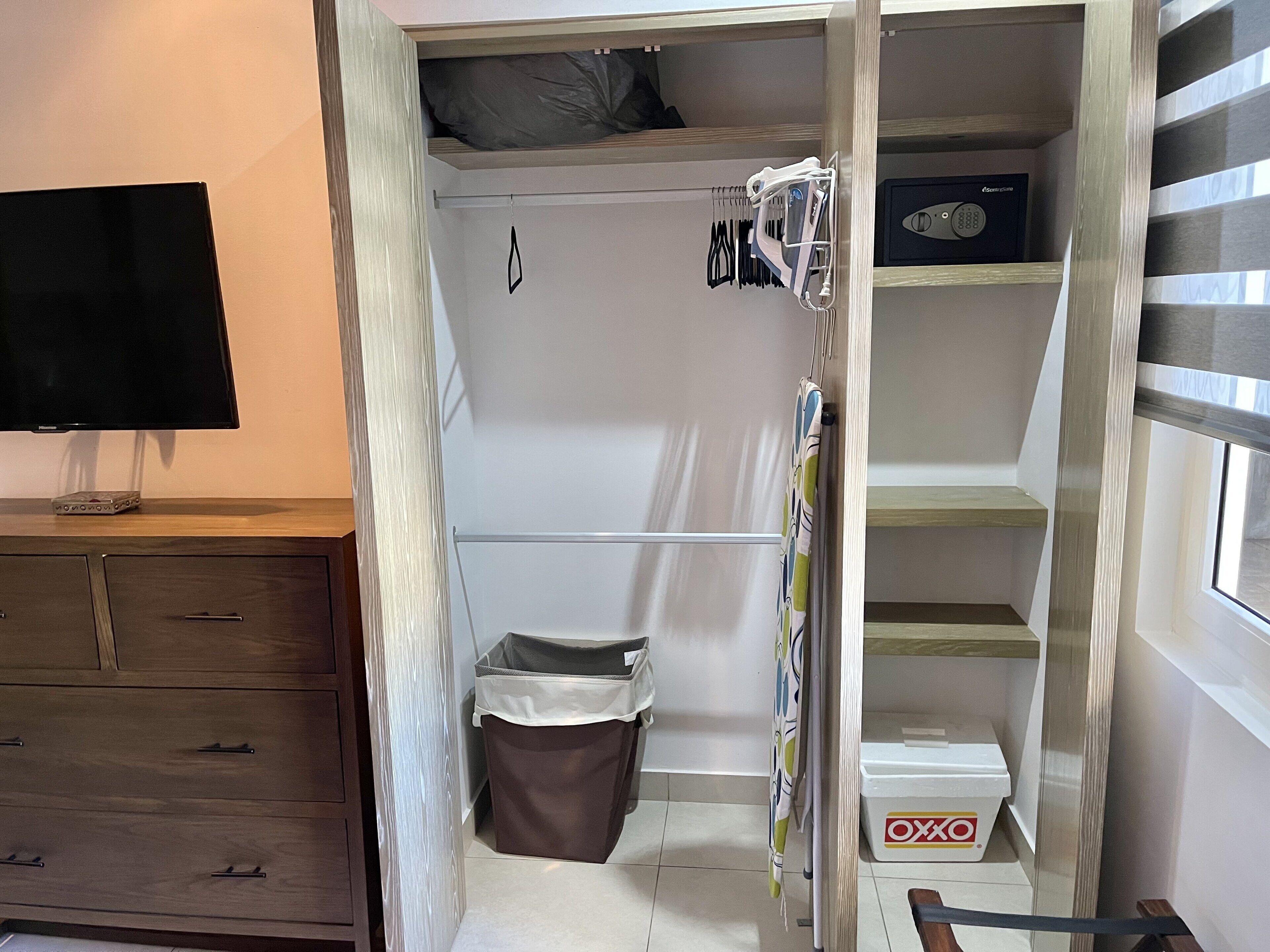 A compact room with a built-in TV, a wardrobe with hanging clothes, a small trash bin, and a window with blinds.