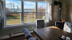 Een knus ingerichte kamer met een groot raam met uitzicht op een meer en bomen, een open haard en een eethoek met een tafel en stoelen.