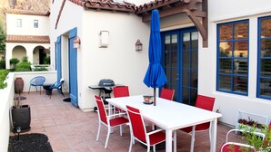A patio with a white table, red chairs, a blue umbrella, and a grill.