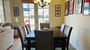A dining area with a table, chairs, and a chandelier. There are framed pictures on the wall and a window with blinds.