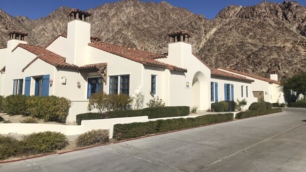 A row of white buildings with red-tiled roofs and blue shutters, set against a mountainous backdrop.
