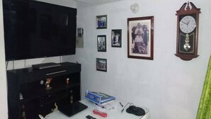 Een woonkamer met een flatscreen televisie, een klok, ingelijste foto’s en een klein tafeltje met een afstandsbediening en een doos.