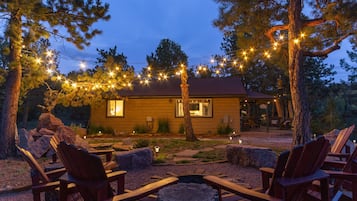 Une cabane chaleureuse avec une terrasse, entourée d’arbres et illuminée par une guirlande lumineuse.