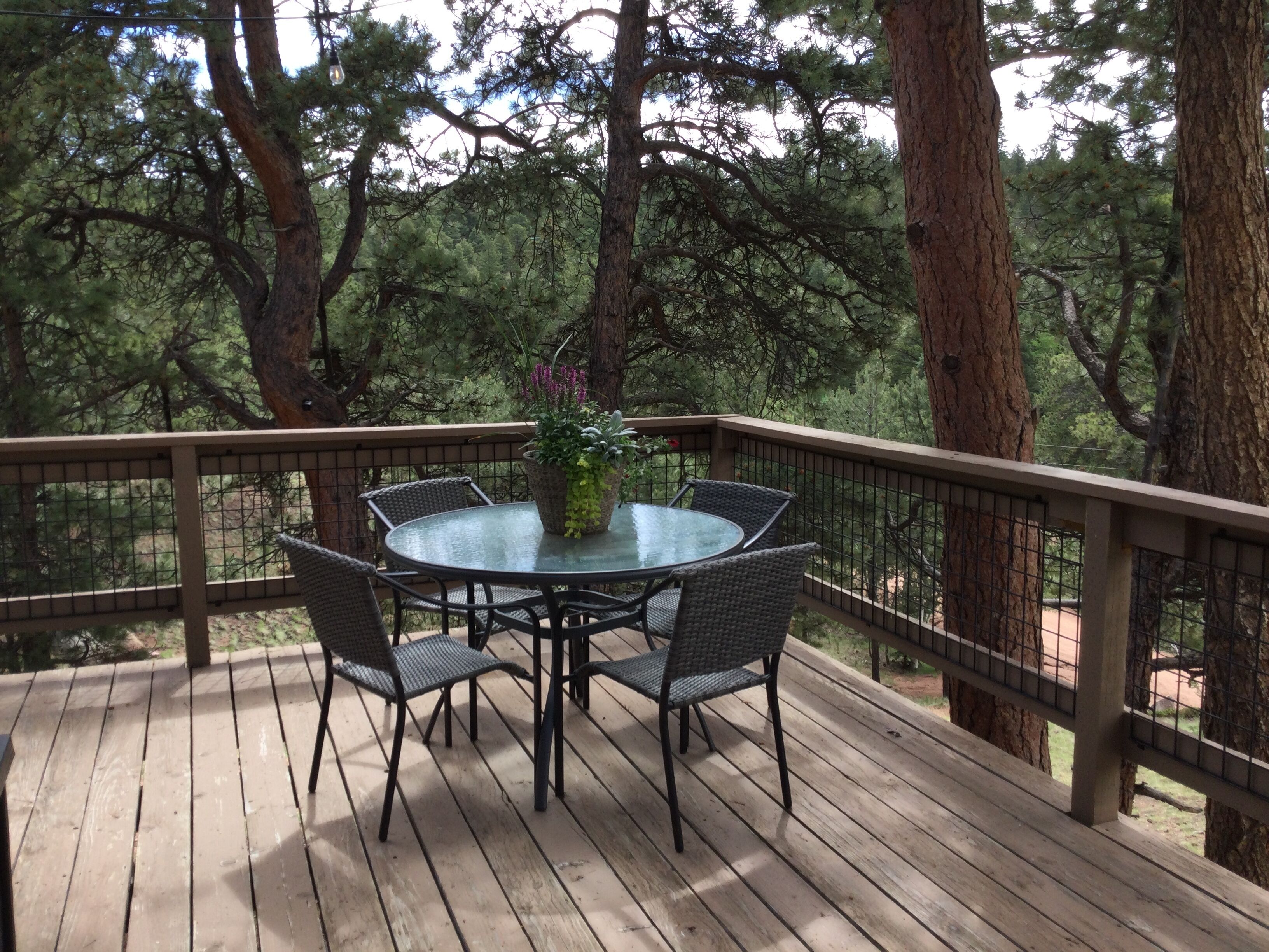 A wooden deck with a glass table and wicker chairs, surrounded by trees.