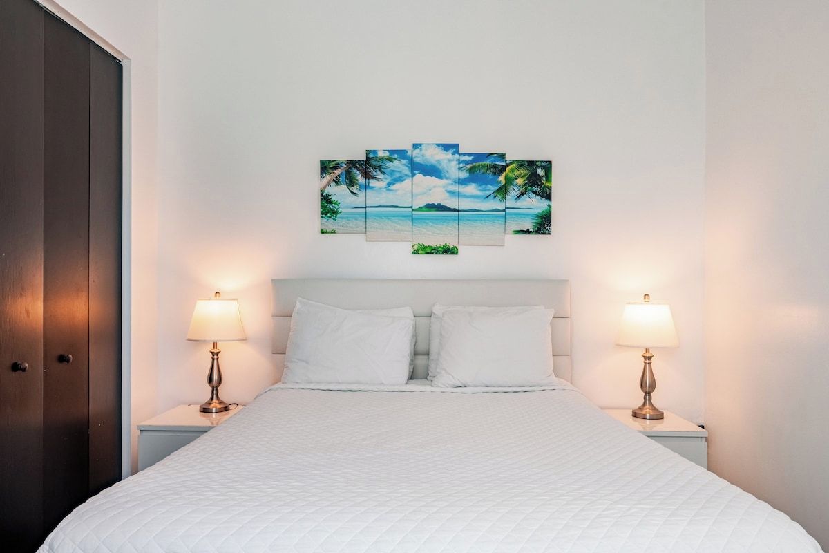 Une chambre d’hôtel avec un lit, deux lampes de chevet, une tête de lit blanche et une œuvre d’art accrochée au mur représentant une scène de plage.