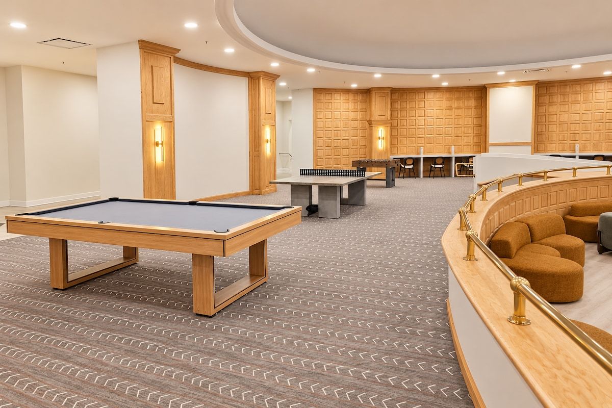 Ein Billardtisch, ein Tischfußballspiel und eine Sitzecke mit gebogener Geländer in einem Raum mit gemustertem Teppich und Holzpaneelen.
