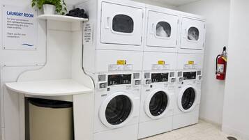 Une buanderie équipée de plusieurs machines à laver, d’une poubelle, d’une étagère avec une plante et d’un extincteur.