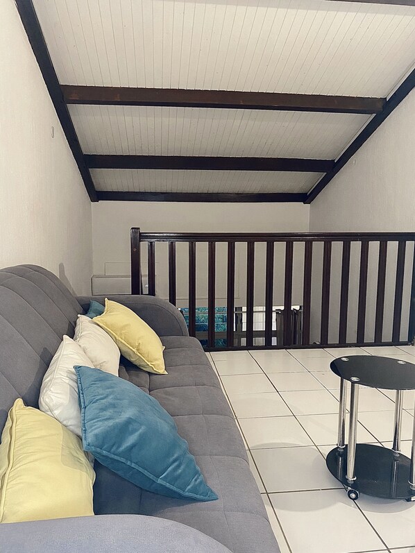 A covered patio area with a grey sofa, multiple cushions, and a small table with wheels.